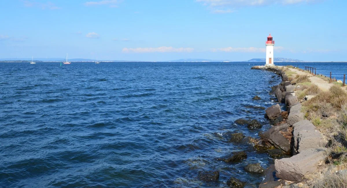 A serene coastal scene featuring a lighthouse at the end of a rocky jetty under a bright blue sky.