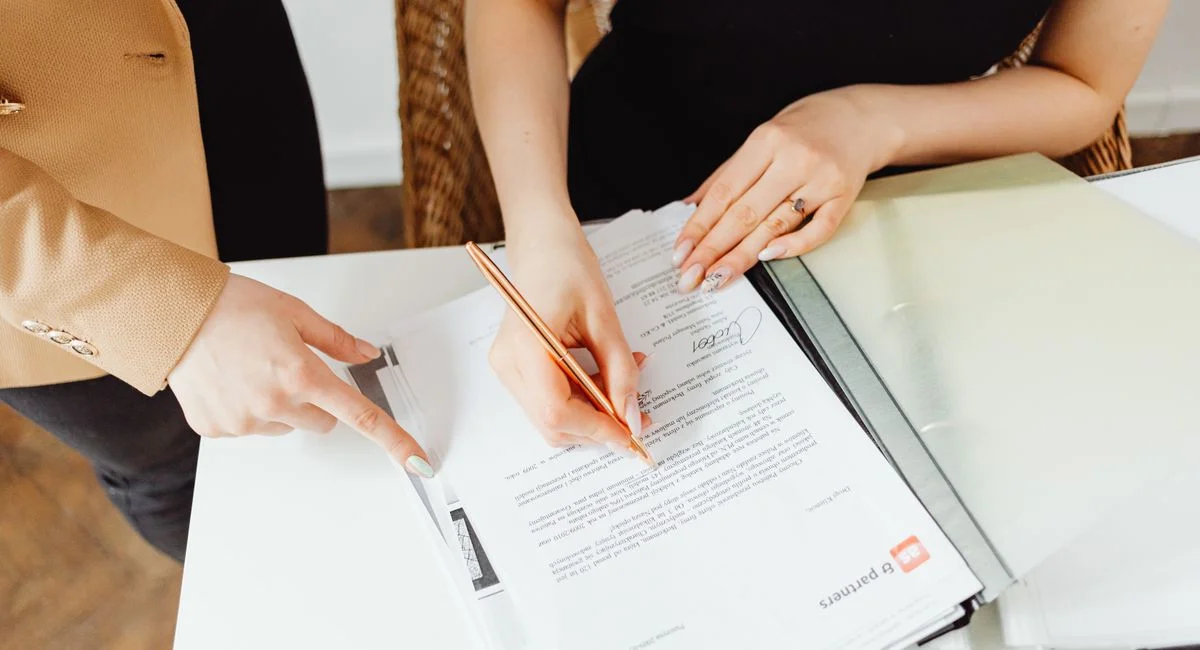 Two women signing important documents at an office desk, showcasing teamwork.