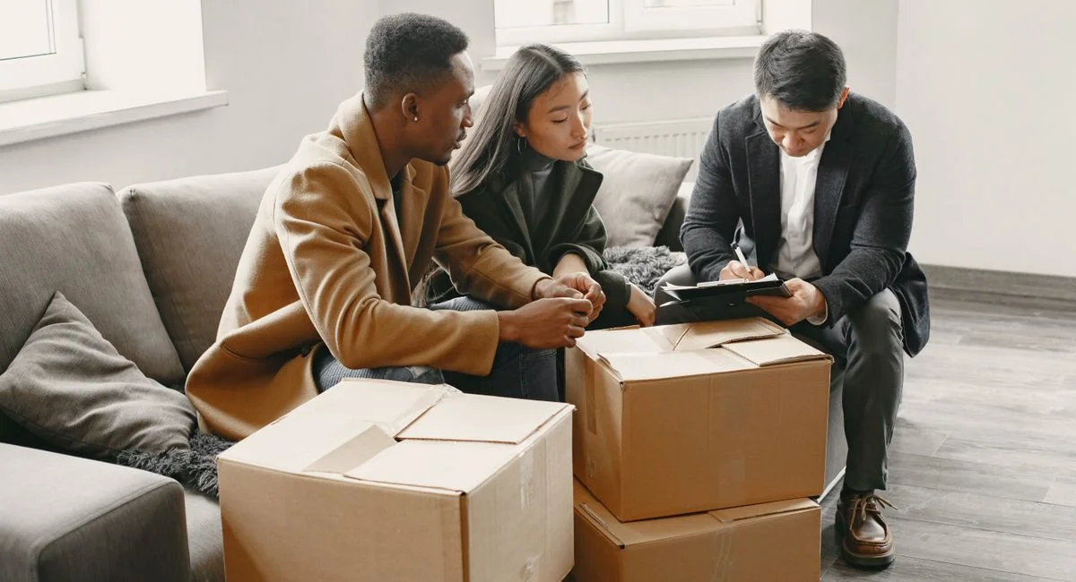 Multiracial couple signing real estate papers at home with a realtor. Cardboard boxes suggest moving in.