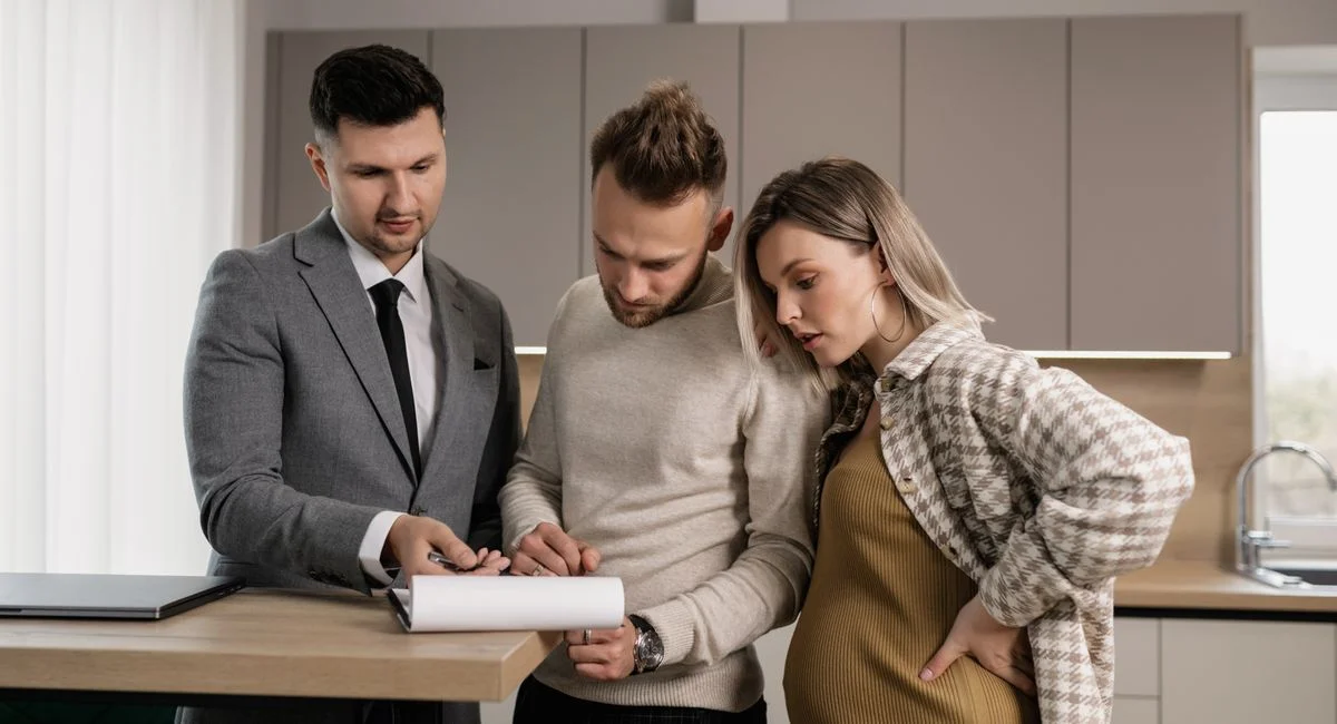 A young couple reviews and signs real estate documents with an agent in a modern kitchen setting.