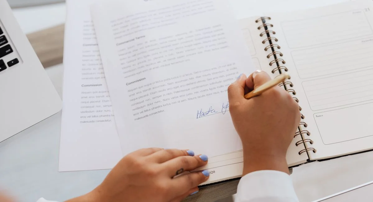 Close-up of hands signing a contract on a desk with office supplies, symbolizing legal agreements.