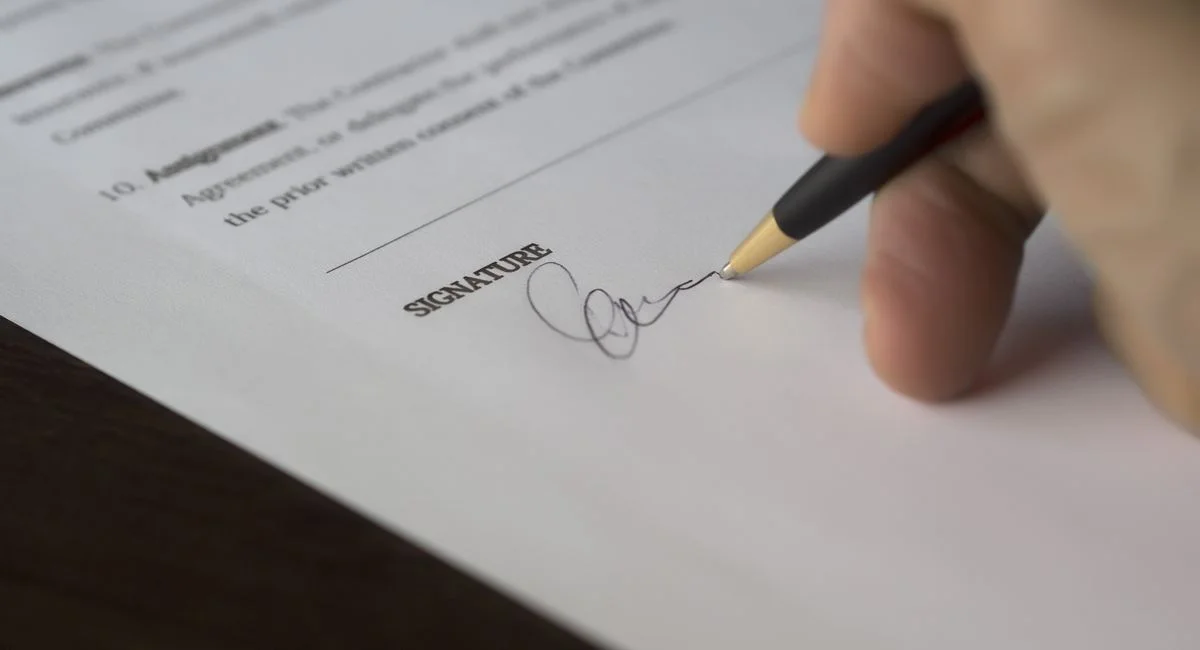A hand signs a formal contract with a pen on a wooden desk.