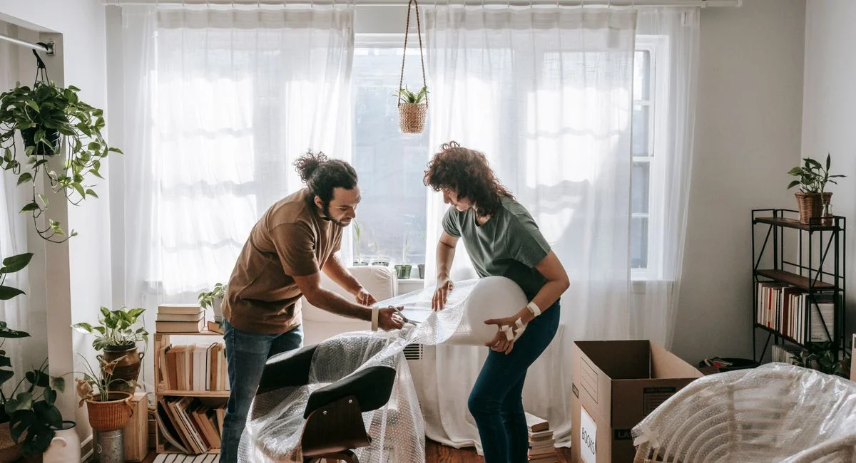 A couple joyfully packing furniture with bubble wrap in a bright, modern living room.