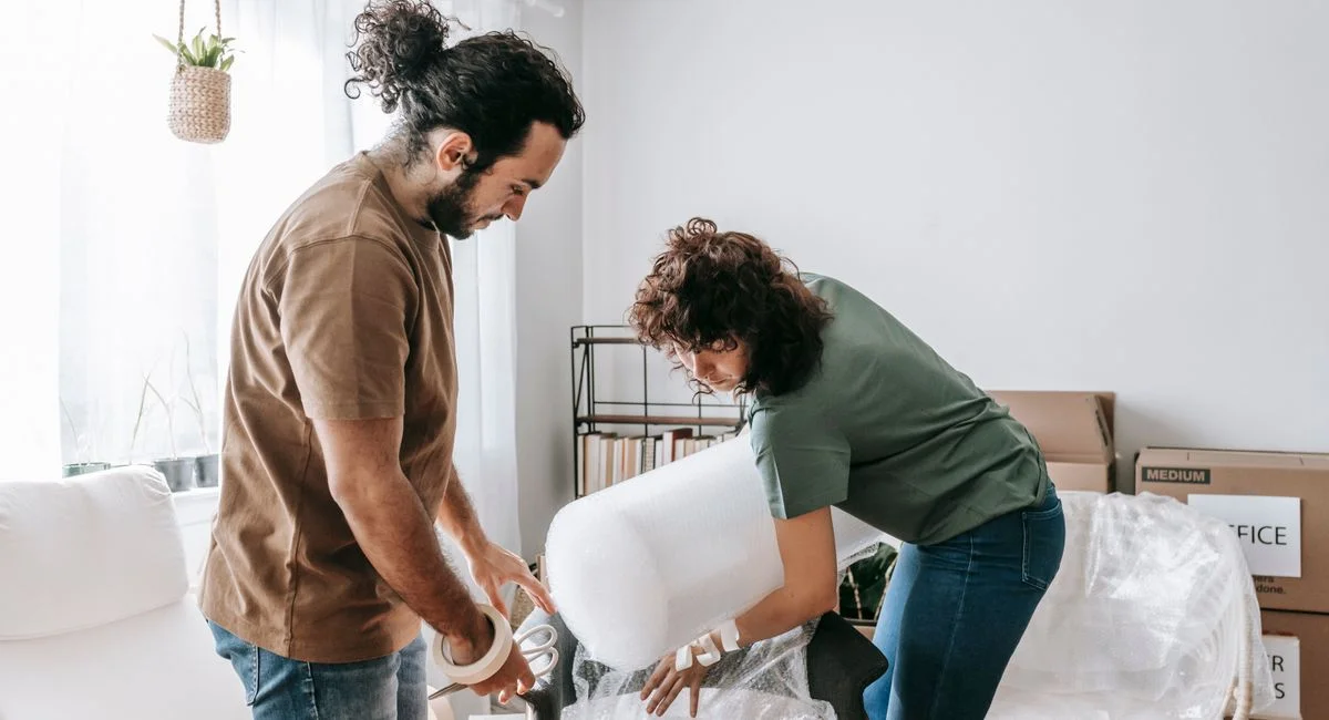 A couple wraps furniture with bubble wrap in a bright and modern apartment while preparing to move.