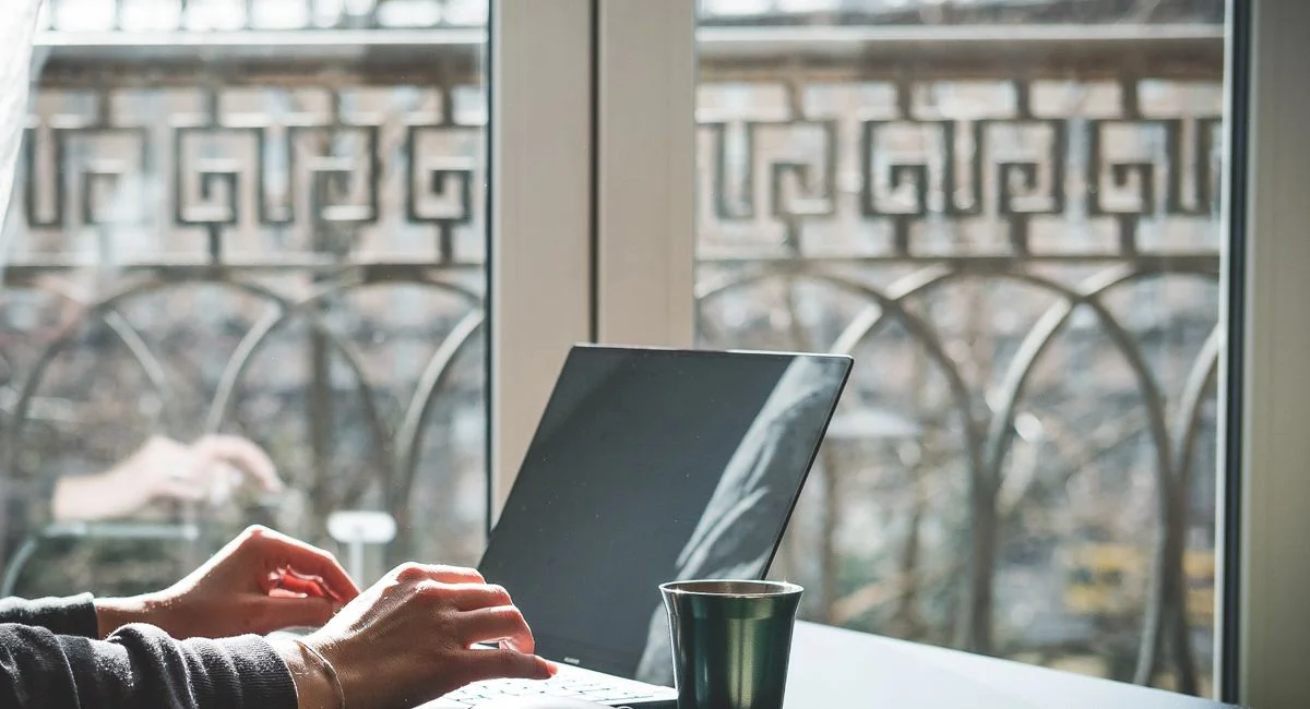 Person typing on laptop at bright workspace with outdoor view, enjoying coffee.