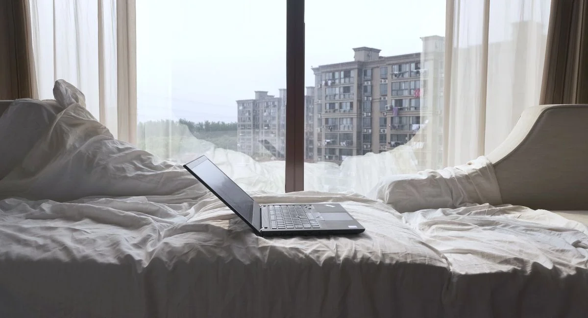 Open laptop on a bed with a view of modern buildings through a Shanghai window.