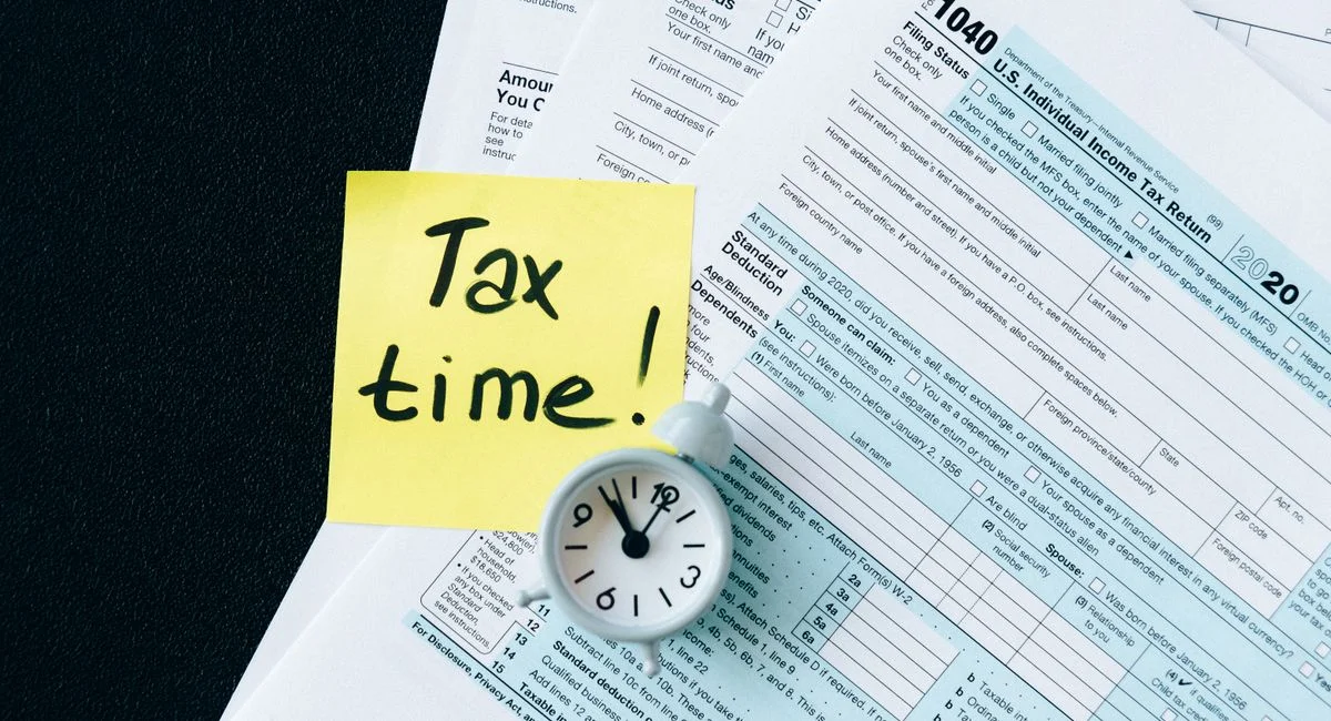 A stack of tax forms with a clock and yellow sticky note saying 'Tax time!' indicating urgency.