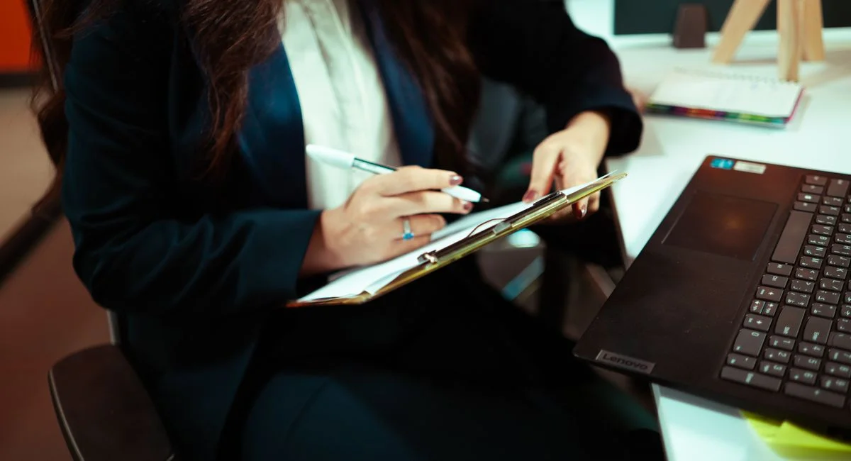 A focused businesswoman writing on a clipboard with a laptop nearby in an office environment.