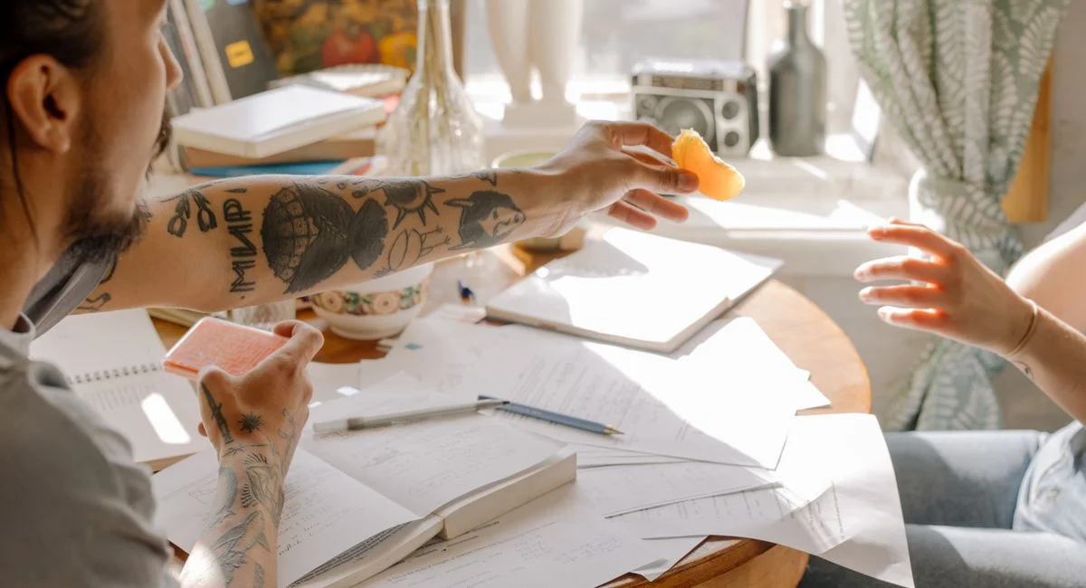 Tattooed students exchanging notes during a study session with papers spread on a round table.