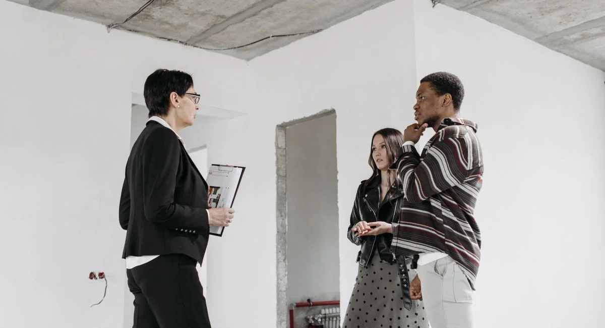 A real estate agent discusses a house plan with a couple in an unfinished room.