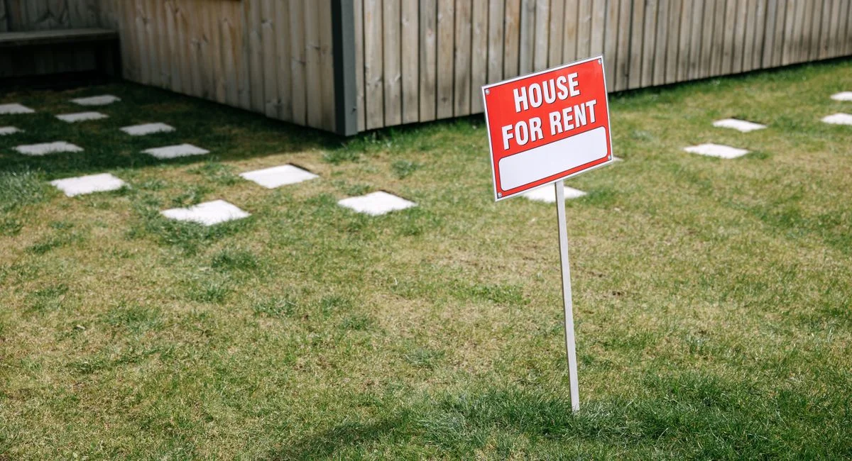 A red 'House for Rent' sign stands on a grassy lawn beside a wooden house exterior.