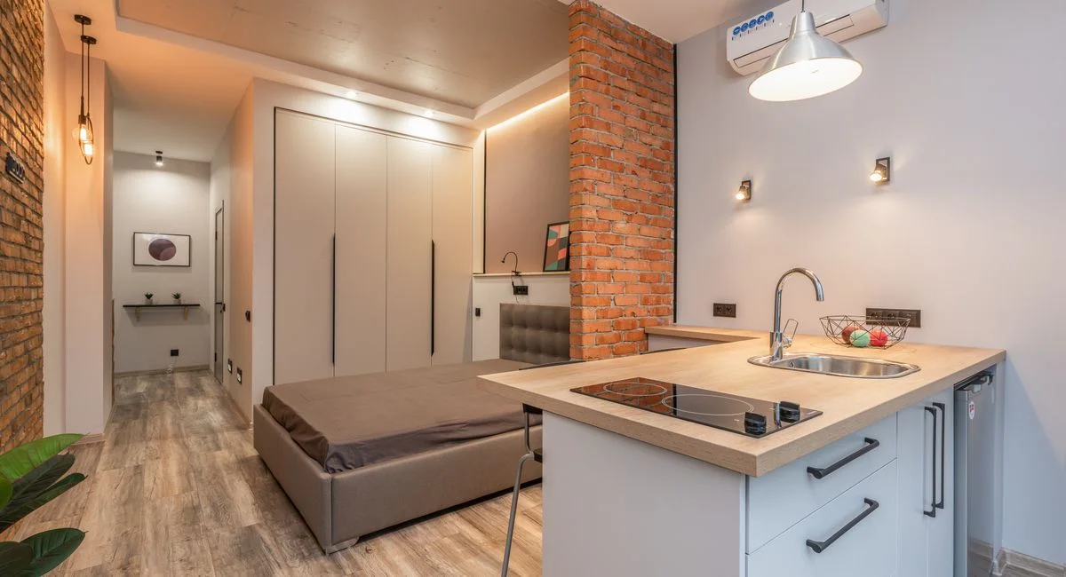 Miniature kitchen with modern appliances and bedroom behind brick partition in small loft style apartment