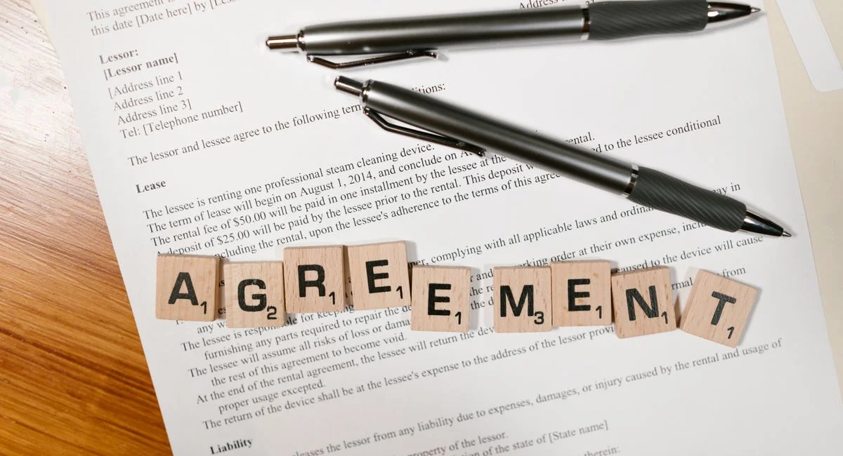 A document with Scrabble tiles spelling 'AGREEMENT' and two pens, symbolizing contract signing.