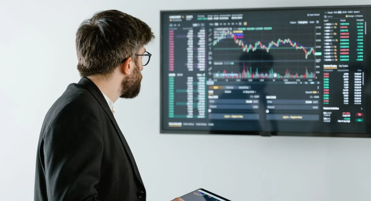 Man observing stock market data on a screen and holding a tablet, representing financial analysis and trading.